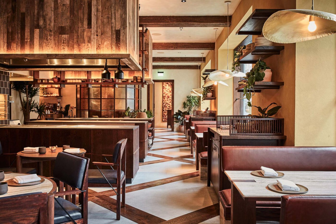 A-nrd-studio-KOL-21 (1) A warmly lit restaurant with wooden accents, leather booths, and tables set with plates and napkins. Hanging lights and plants add a cosy atmosphere.