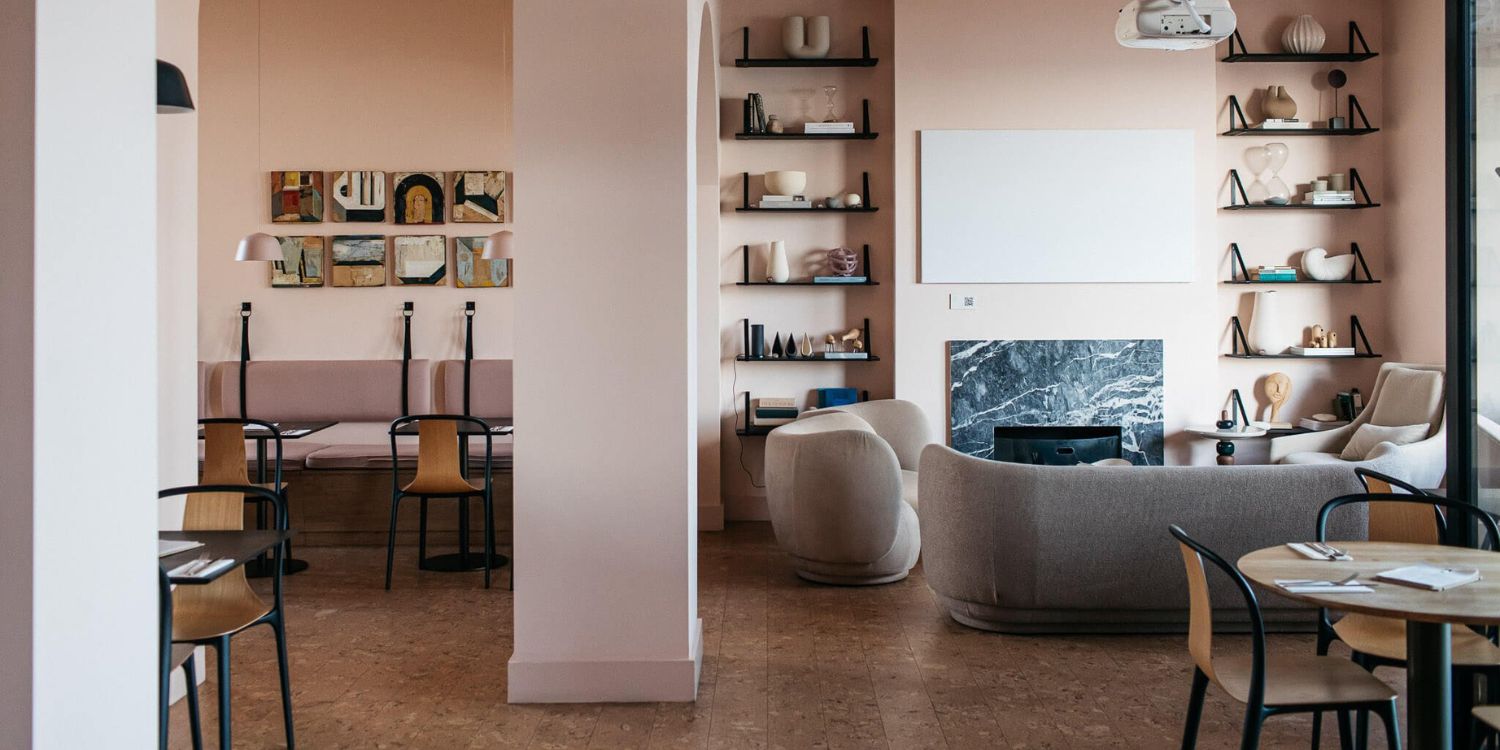 A minimalist living room with soft pink walls features a modern beige sofa, round chairs, and a cosy fireplace. Shelves display decorative items, and a dining area with a table and chairs is visible through an archway.