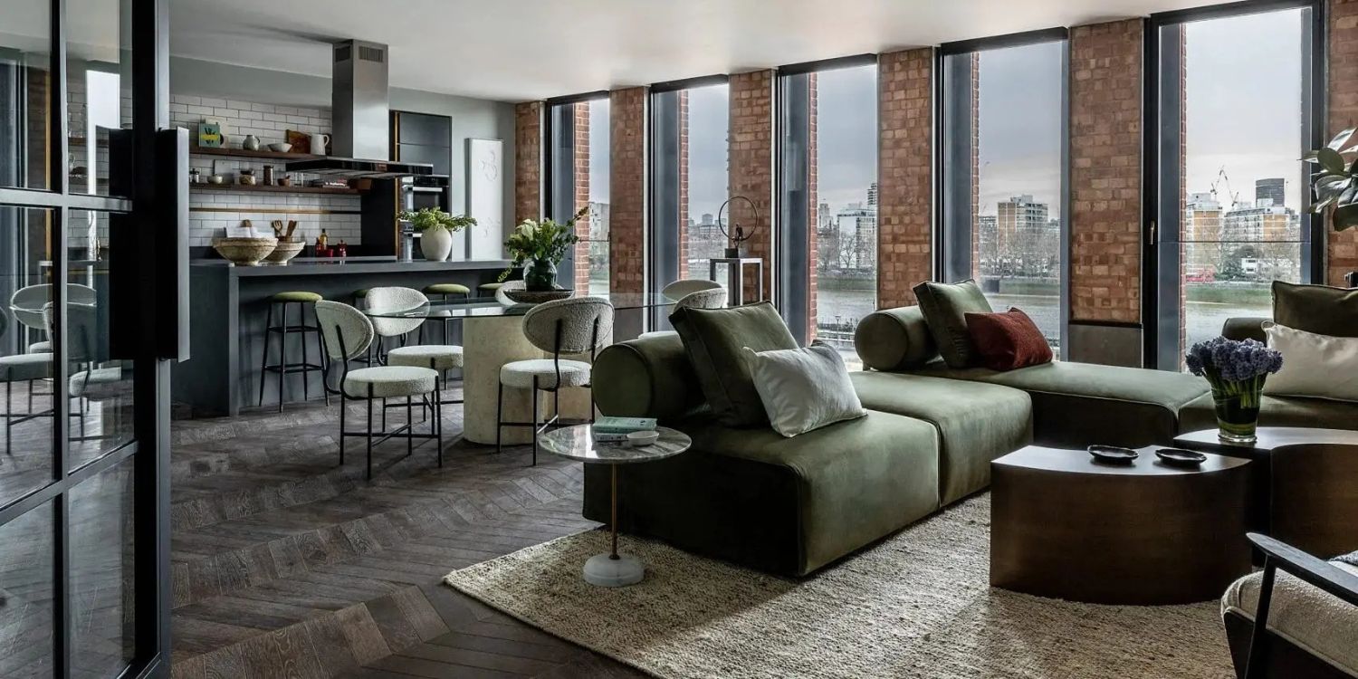 A modern living area with large windows showcasing city views. The room features green sofas, a round coffee table, and a kitchen with bar stools. Wooden and brick accents add warmth to the space. A vase of flowers decorates the table. A perfect example of industrial interior design.