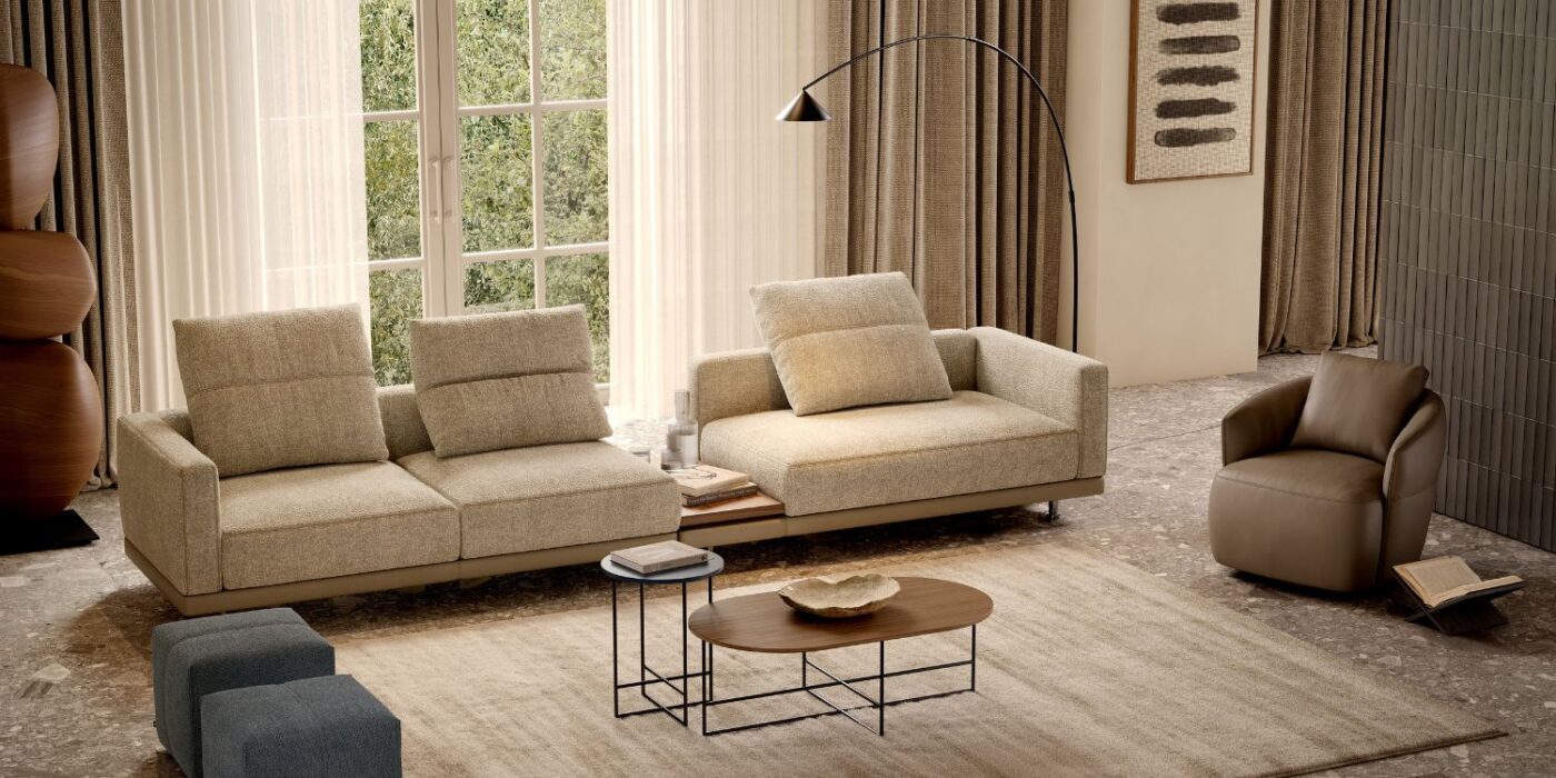 Blog Featured Image - 2026-03-10T165829.951 A cosy living room with neutral tones, featuring a beige sectional sofa, a brown armchair, a round wooden coffee table, and floor lamp. Large windows with sheer curtains let in natural light, creating a warm, inviting atmosphere. Danish interior design