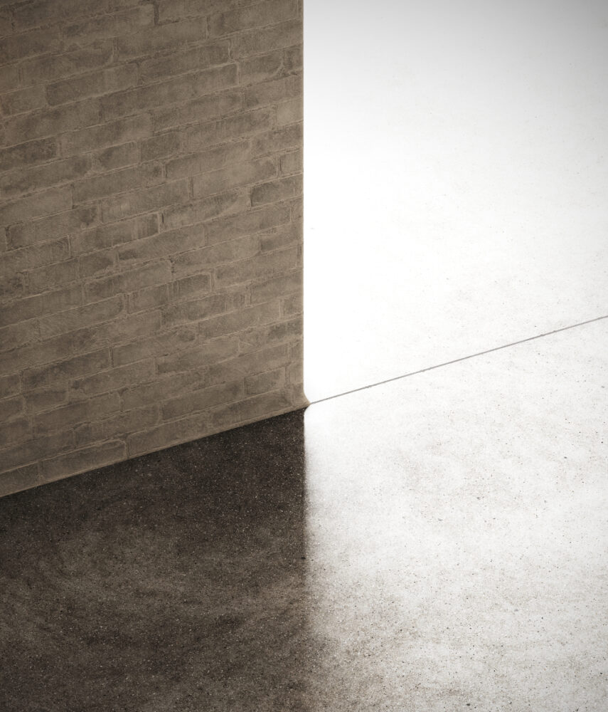 A tall brick wall divides two types of flooring: a dark, textured surface on the left and a light, smooth surface on the right. Bright light casts an angled shadow on the ground, emphasizing the contrast between the materials.