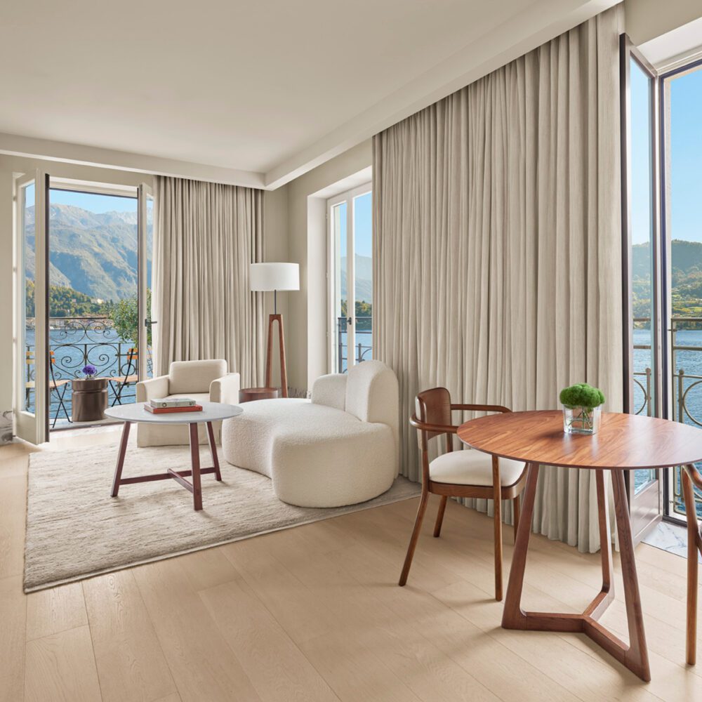Como1_01b_524_RGB_V1 by edition hotels Bright living room with modern white and wood furniture, large windows, mountain and lake view. Elegant, serene, inviting ambiance.