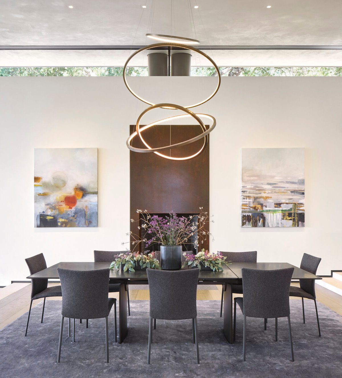 Forest Aerie Project by Shawback Design. The dining room is in white and grey tones with some gold accents in the lighting. It has a wood dining table with six dining chairs around it. In the middle of the dining table it has a vase with flowers.