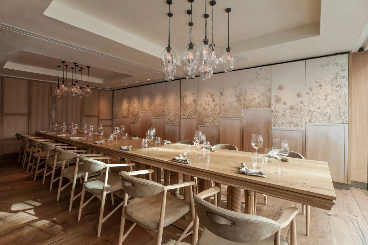 Hide-Restaurant-Dabbous-Interior-Designer-Dining-Room-Above-TWW-web Elegant dining room with a long wooden table set for a meal. Soft lighting from modern hanging fixtures and floral wall panels create a cosy atmosphere.