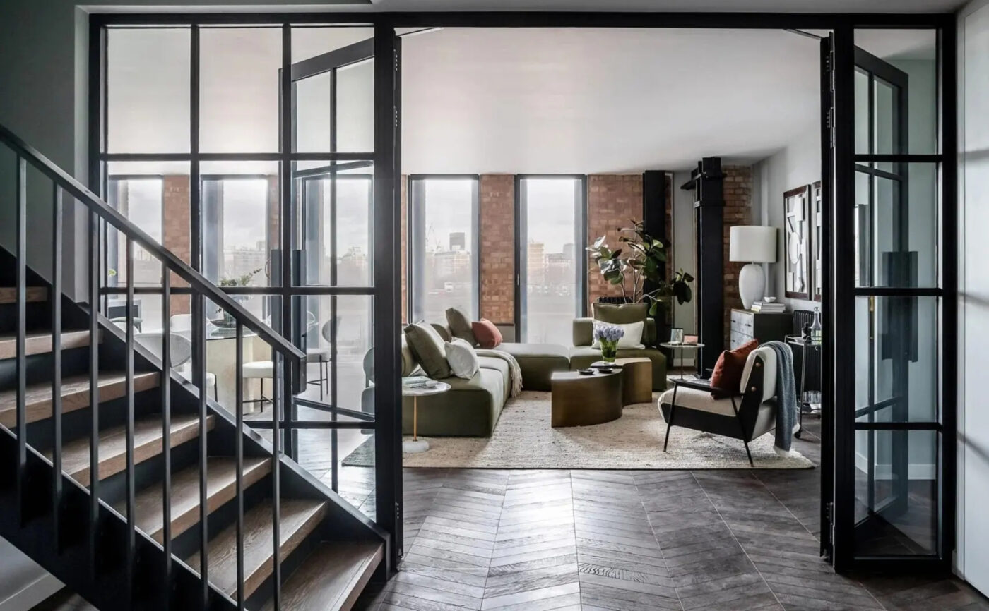 A modern living area with large windows showcasing city views. This industrial interior design room features green sofas, a round coffee table, and a kitchen with bar stools. Wooden and brick accents add warmth to the space.