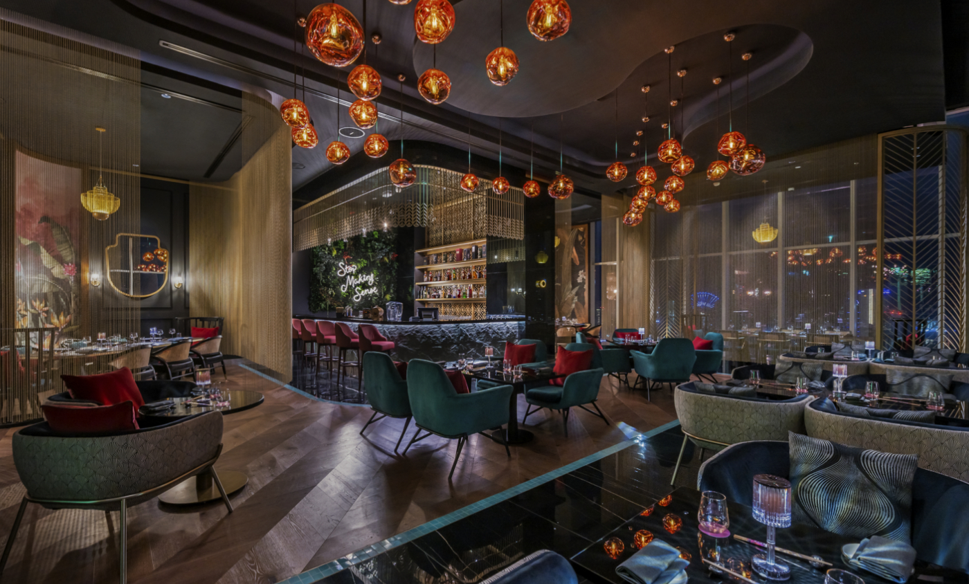 Restaurant design in dark tones. The chairs are in dark green and others with a floral pattern. The lights are orange and suspended in the ceiling. With a bar in the corner with red bar chairs.