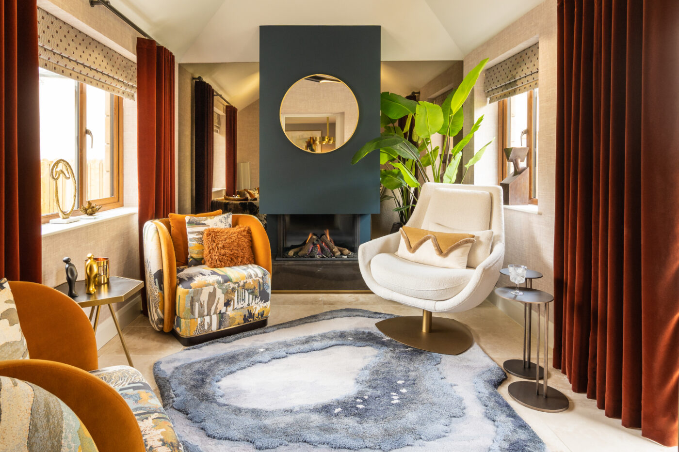 A cosy living room with a modern fireplace, round mirror above it, and large green plant beside it. The space includes a white swivel chair, patterned armchairs, and a textured rug in shades of blue and gray, with large windows draped in burgundy curtains.