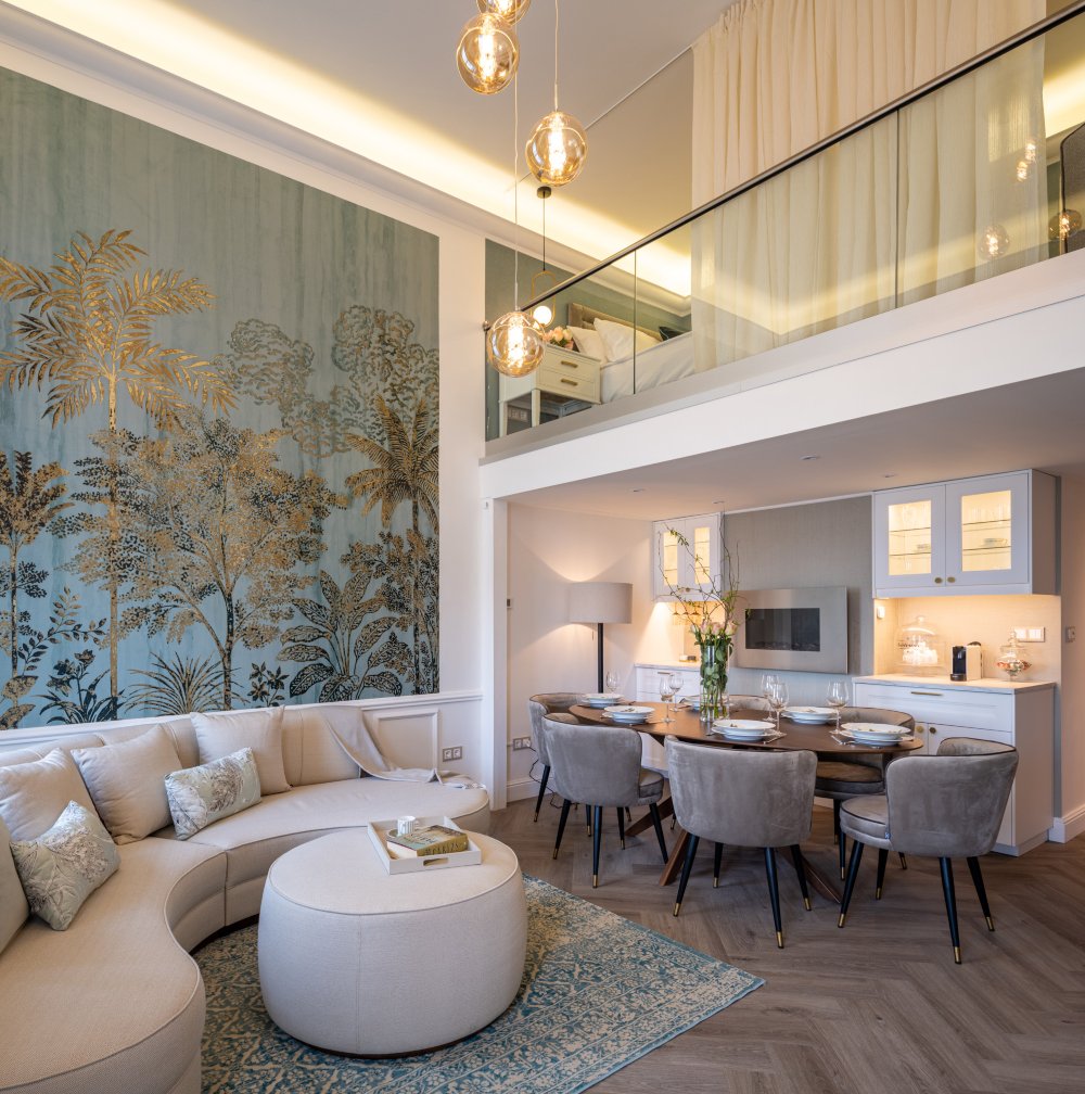 Living room and dining room with a white sofa, white pouf, oval dining table and six brown dining chairs. With a wall with a blue print and gold accents.