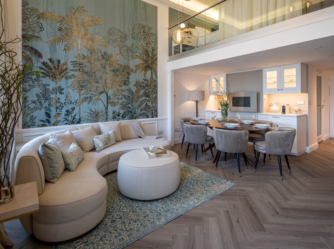 Living room and dining room with a white sofa, white pouf, oval dining table and six brown dining chairs. With a wall with a blue print and gold accents.