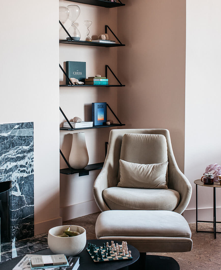 A minimalist living room with soft pink walls features a modern beige sofa, round chairs, and a cosy fireplace. Shelves display decorative items, and a dining area with a table and chairs is visible through an archway. Port Hotel