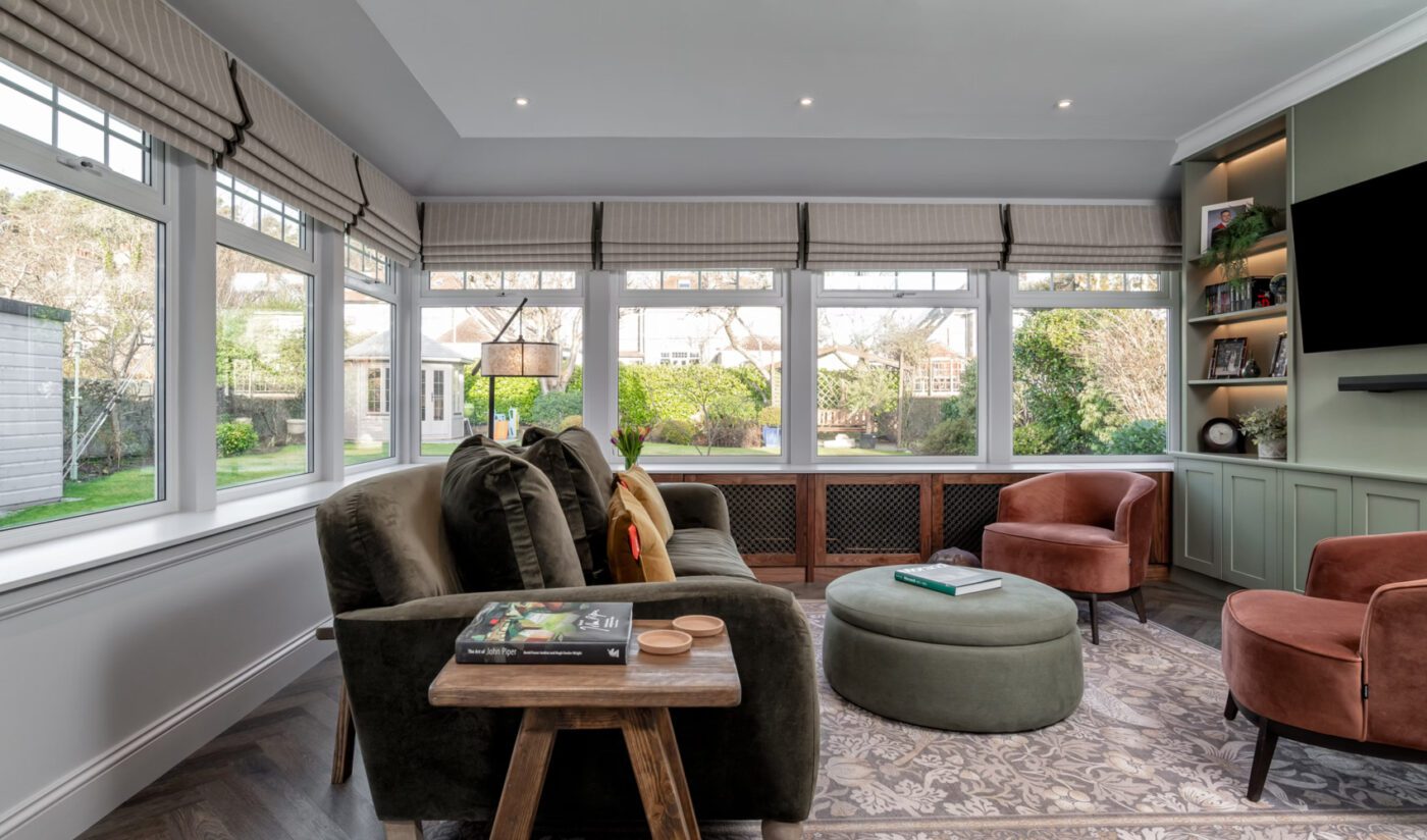 Project-by-Zhanar-Design-Photography-by-Niall-Hastie-Photography-2 A cosy living room with a large window view of a garden. It features olive green and brown seating, a round pouf, a patterned rug, and a wall-mounted TV, conveying comfort and warmth.