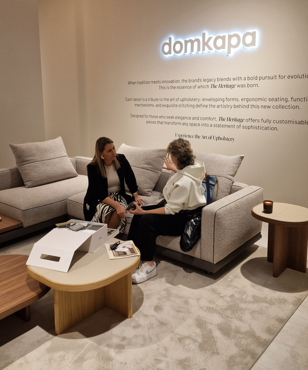 Two women sit comfortably on a sofa, with a "Domkapa" sign visible behind them. They are discussing a project and selection the materials with the help of a sample box.