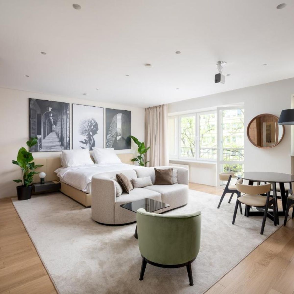 Specifying Upholstery For Hotel Suites (1) A modern, airy hotel suite features a bed with white linens, abstract art, and indoor plants. A cozy seating area and dining table create a serene, inviting space.