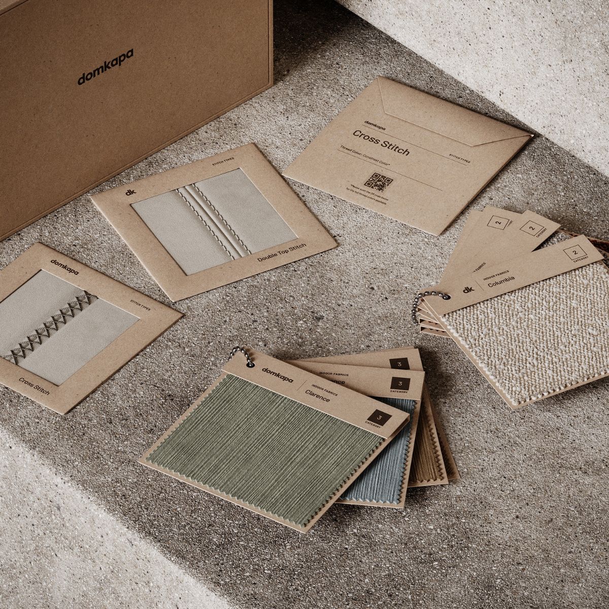 Specifying Upholstery For Hotel Suites (3) Fabric swatches and brown envelopes with stitching patterns labeled "Cross Stitch" on a textured concrete surface, conveying a neutral, creative tone.