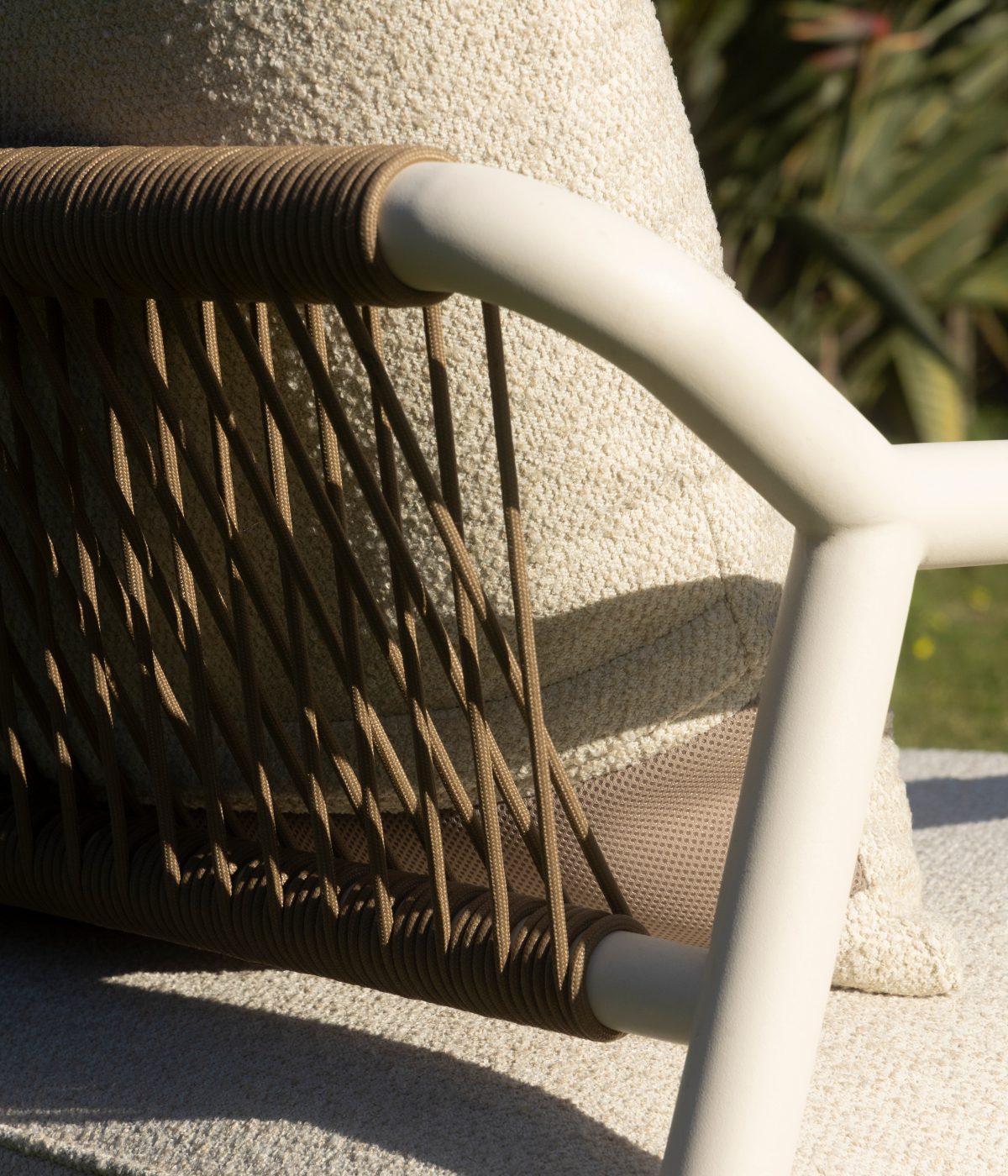domkapa_campaign_@11artfactory-074_usada Close-up of a modern outdoor chair with light tan woven ropes and a cream frame. Soft sunlight highlights the texture of the fabric and the greenery in the background.