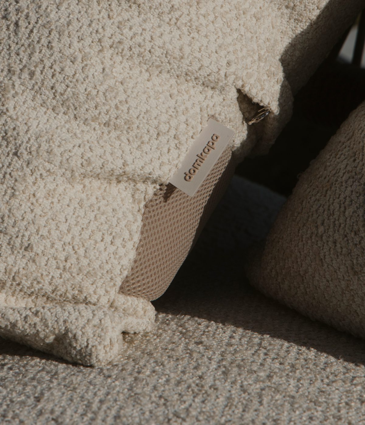 domkapa_campaign_@11artfactory-079 Close-up of a textured cream pillow with a small fabric tag, placed on a woven surface. The scene conveys warmth and comfort in soft lighting.