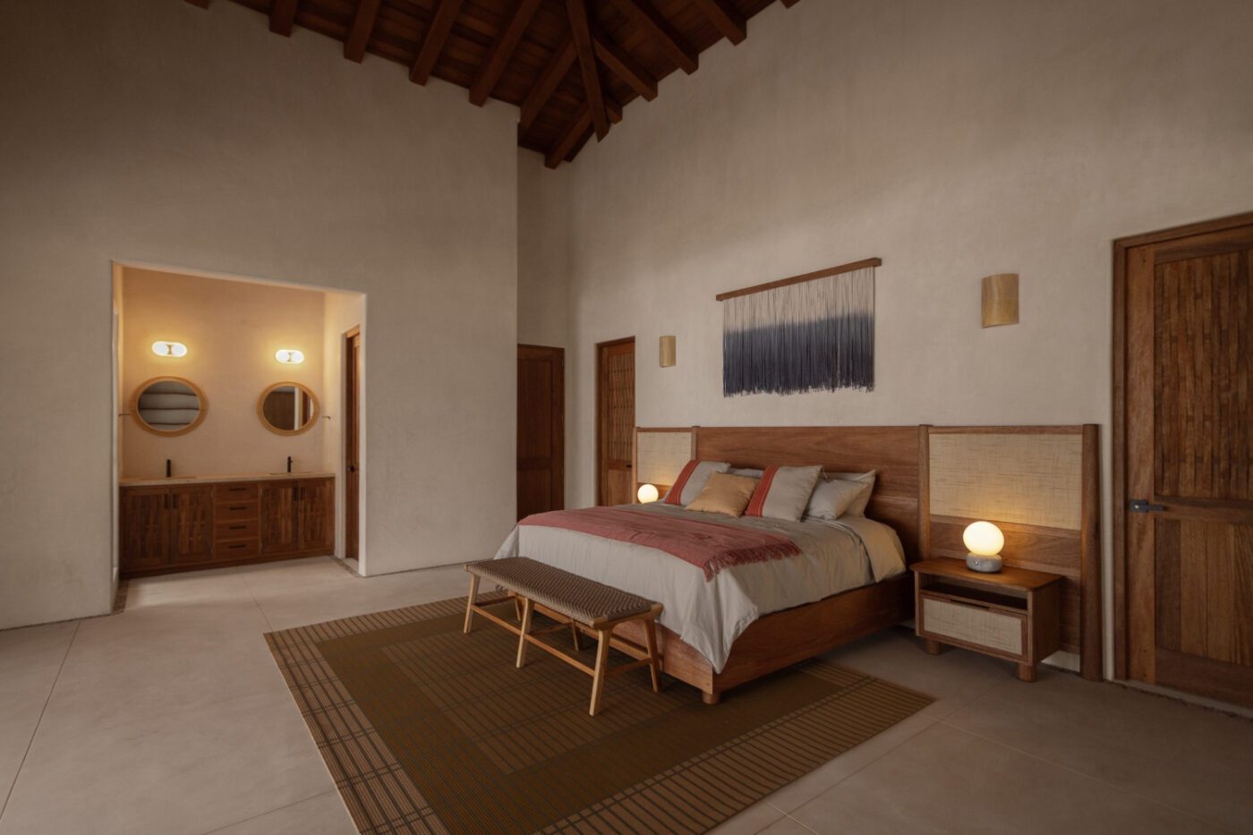 imgi_43_CPMX—Troncones—09-scaled Cosy bedroom with high ceiling, wooden bed and earthy tones. Soft lighting, two doors, a woven wall hanging and a glimpse of a bathroom are visible. By one of the best mexican interior designers.