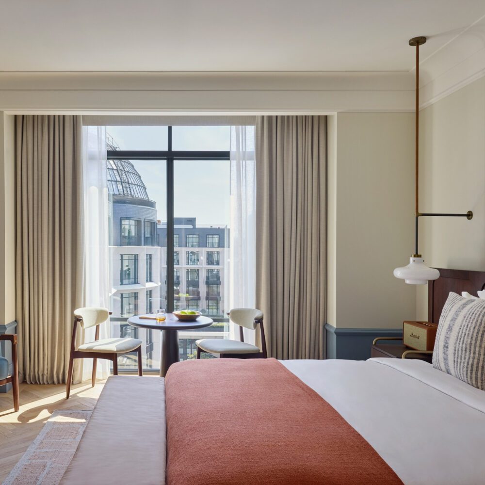 imgi_88_Premier___Premier_Terrace_View by six senses Spacious modern hotel room with a large bed, orange throw, glass enclosed shower, wooden armchairs, and floor-to-ceiling window with city view.