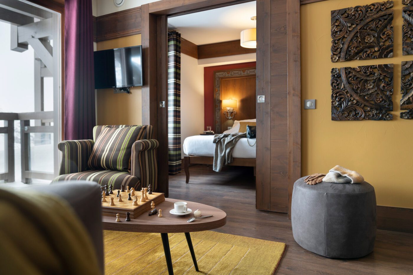 kashmir-suite-junior-n105-studiobergoend A modern hotel suite with a striped armchair, chessboard on a coffee table, tea set, and a view into a bedroom with a neatly made bed and decorative wall art.