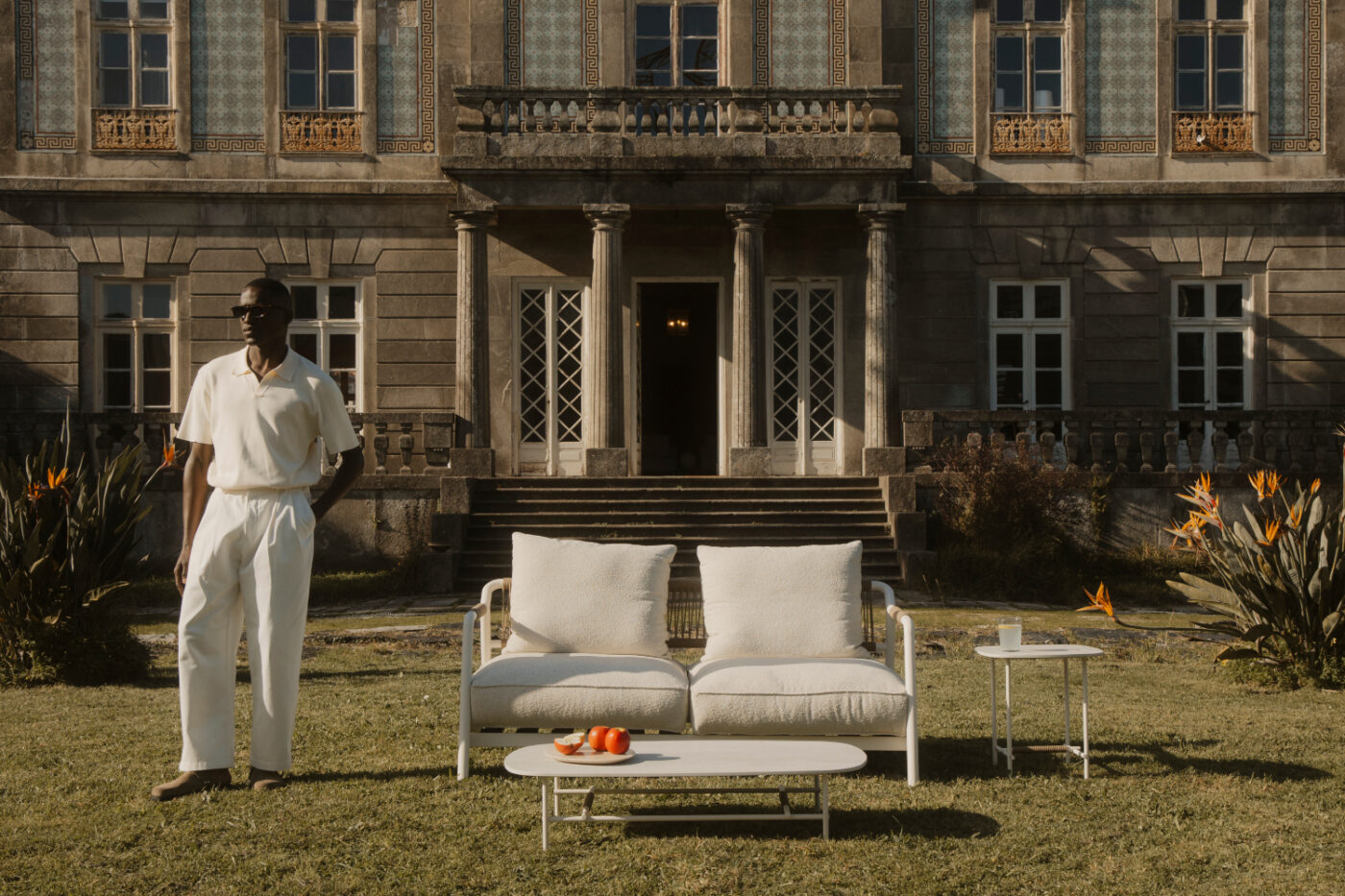 A man dressed in white poses in front of a grand mansion, highlighting its size and beauty. An outdoor collection is by its side. Outdoor furniture care in sunny days.