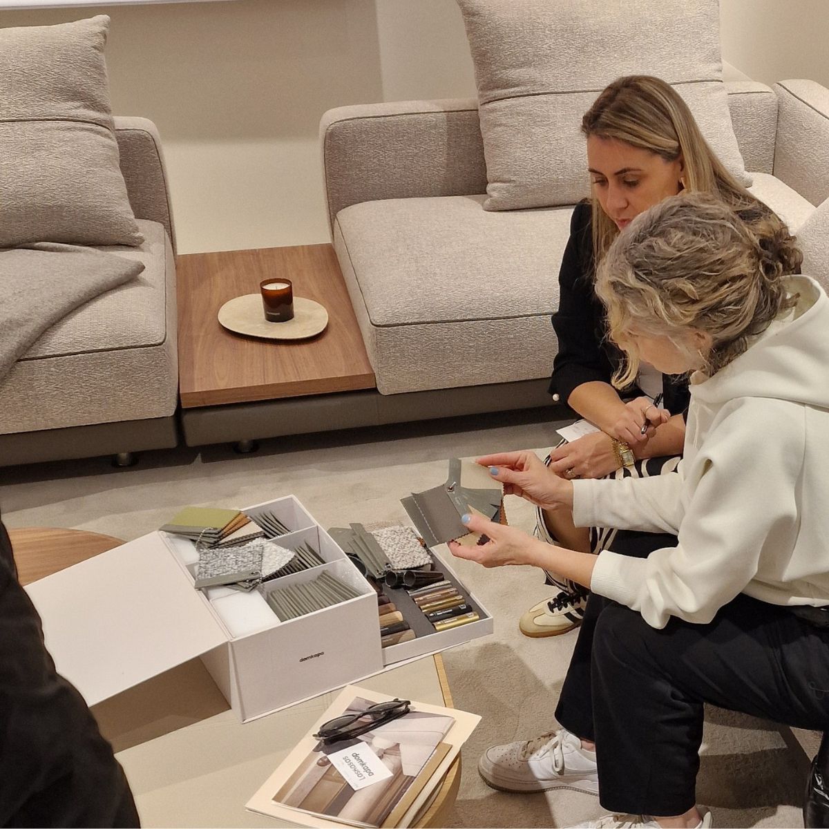 upholstery grades by domkapa Two women are seated on a sofa, focused on a sample box in front of them, sharing a moment of interest and engagement.