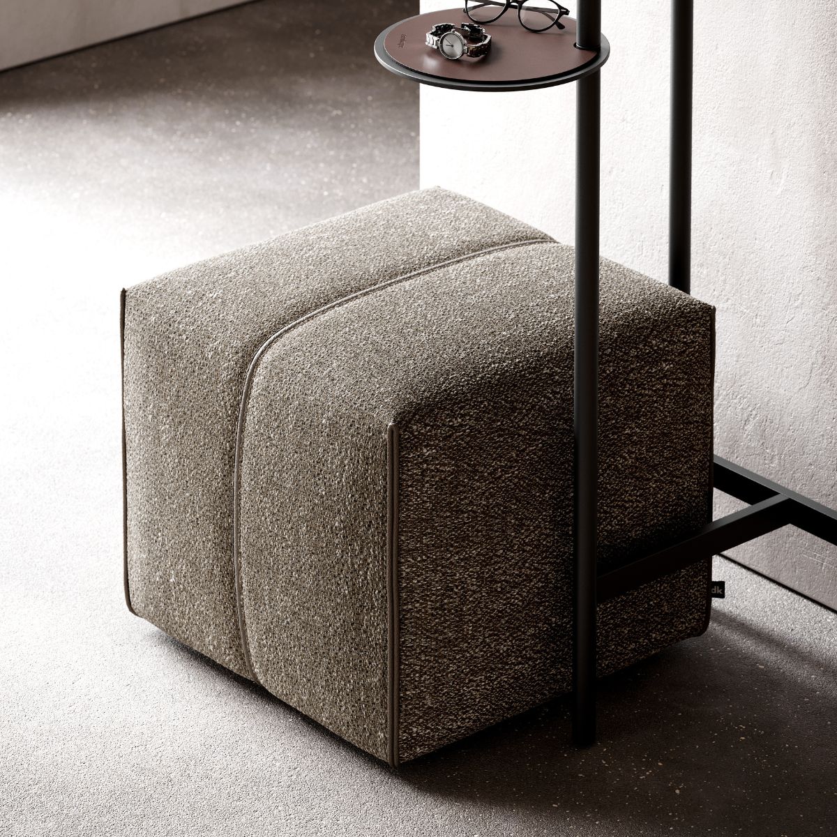 upholstered poufs selection (3) A textured gray pouf sits on a speckled floor next to a slim, black metal stand with a round shelf holding glasses and a watch. The setting feels modern and minimalist.